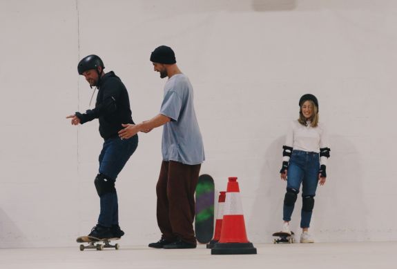 man being helped by an instructor with woman looking on at a skateboard lesson for two