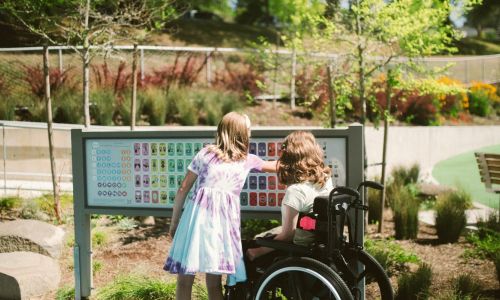 Two young girls one in a wheelchair in a park looking at a sign