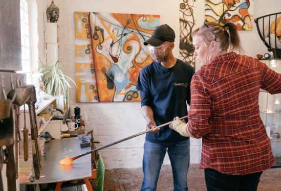 a woman being shown how to do glassblowing in a workshop on a glassblowing experience