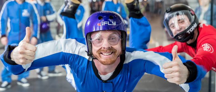 a man wearing a blue flight suit, goggles and helmet giving the thumbs up on an indoor skydiving experience