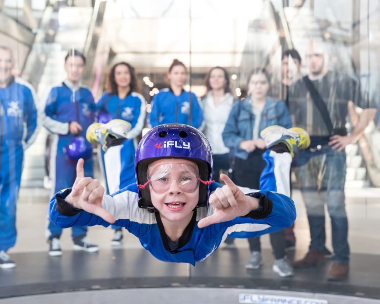 a young child wearing a blue flight suit, goggle and helmet on an indoor skydiving experience being watched by family members