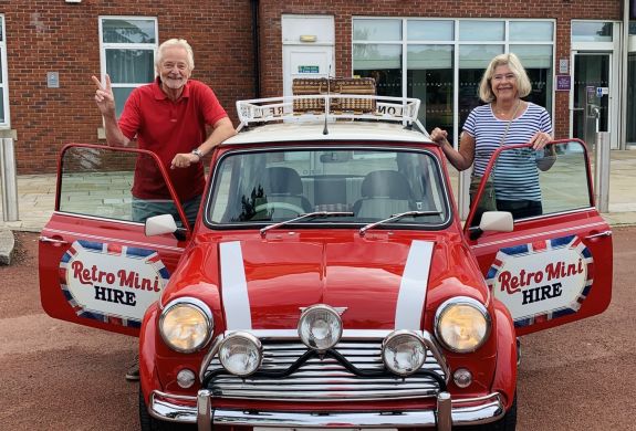 a man and a woman standing either side of a classic red mini cooper on a self drive road trip