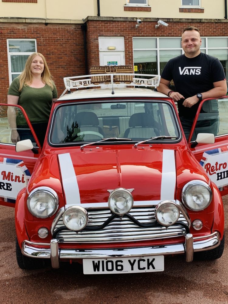 a man and a woman standing either side of a classic red mini cooper on a hire and drive experience day