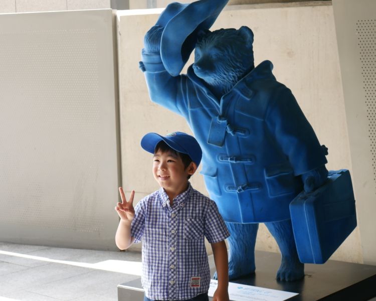 Young boy posing for photo infront of blue statue of paddington bear while on a paddington walking tour in london