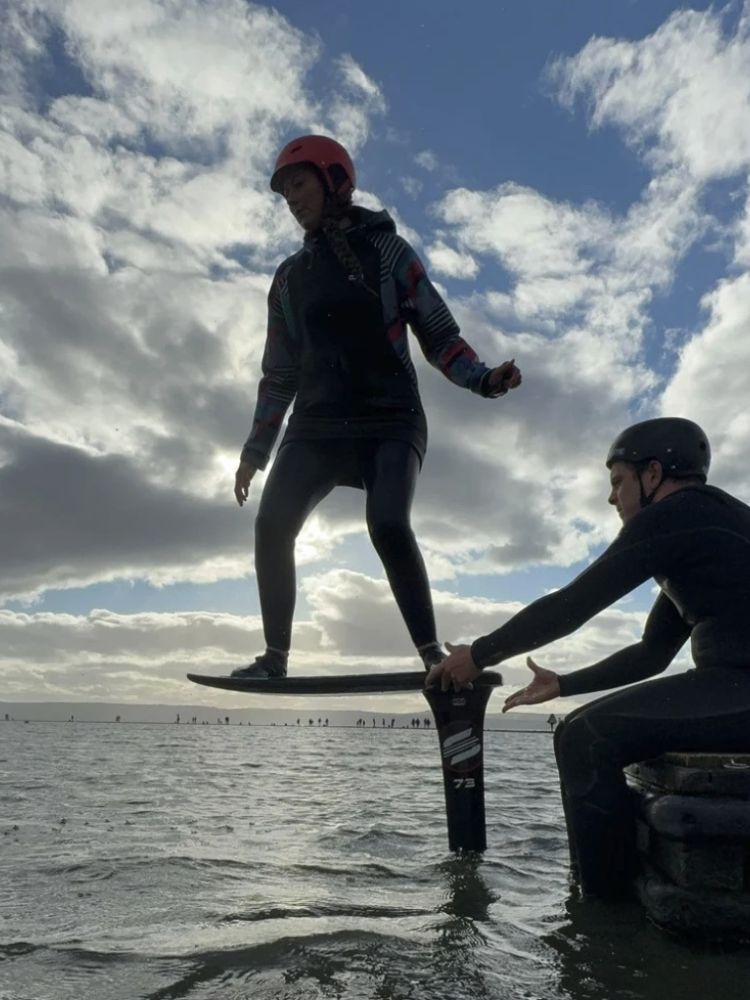 woman standing on a pump foil board with instructor sitting on the side of the water