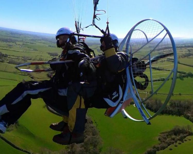 side view of a tandem paramotor experience flying over green fields against blue skies