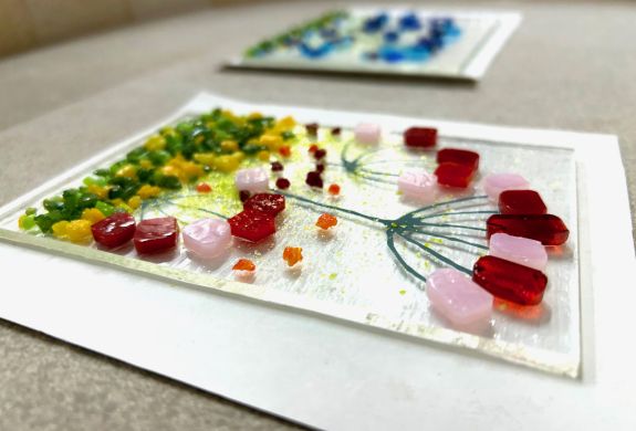 the front of a glass tea light holder with a coloured floral design laid on a board ready to go into the kiln