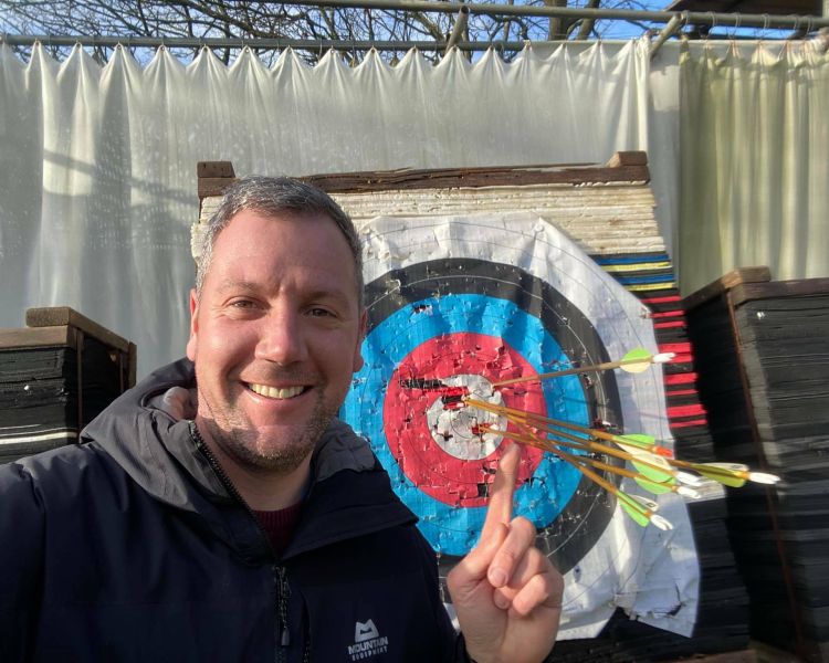 man smiling standing in front of a target full of arrows as part of an archery experience