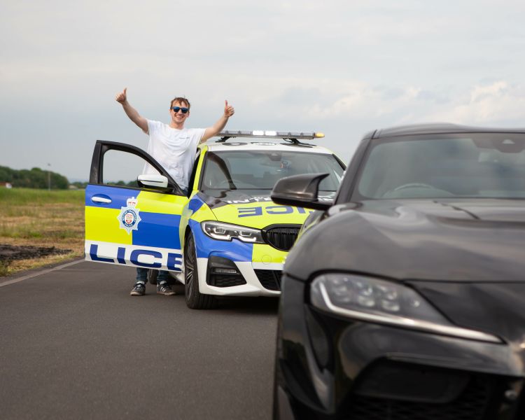 man in white t-shirt wearing sunglasses giving thumbs up next to a police interceptor bmw with a black sports car ahead as part of a police interception driving experience